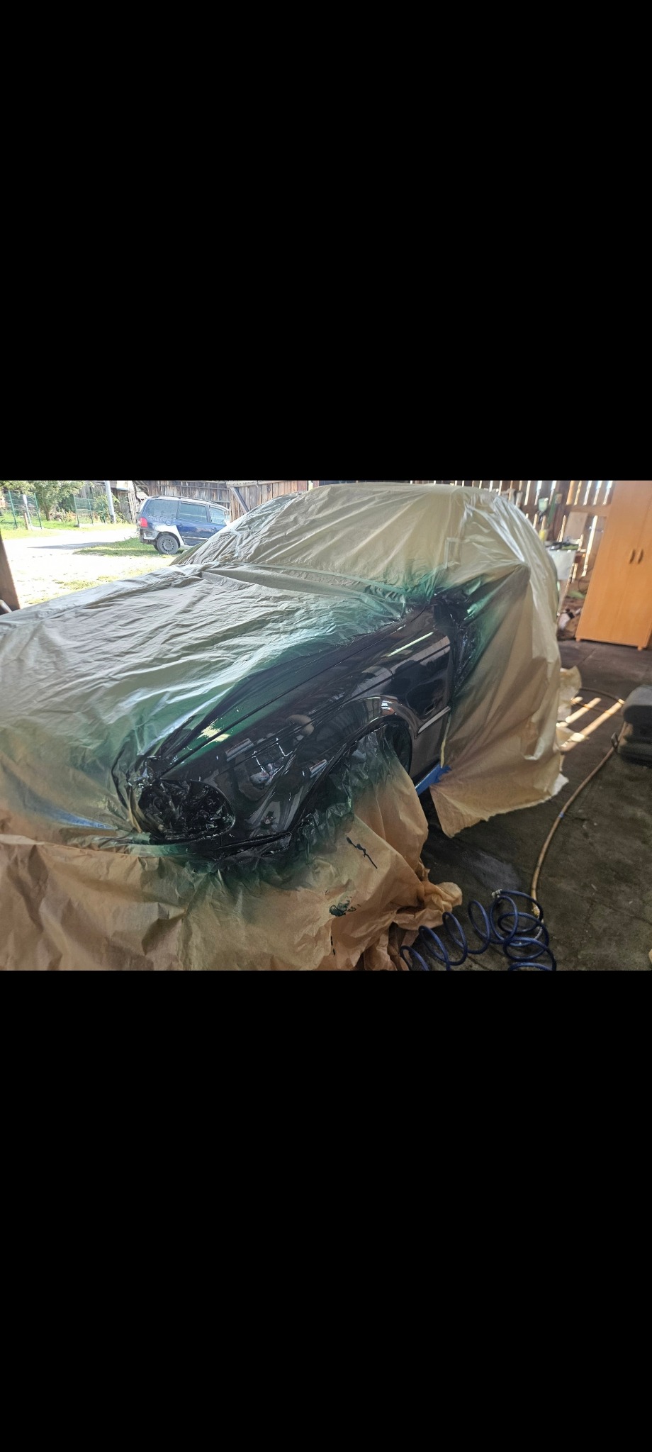 Samochód częściowo zakryty folią ochronną przed lakierowaniem, widoczne ślady farby na folii i elementach auta, niebieski wąż pneumatyczny leży obok.