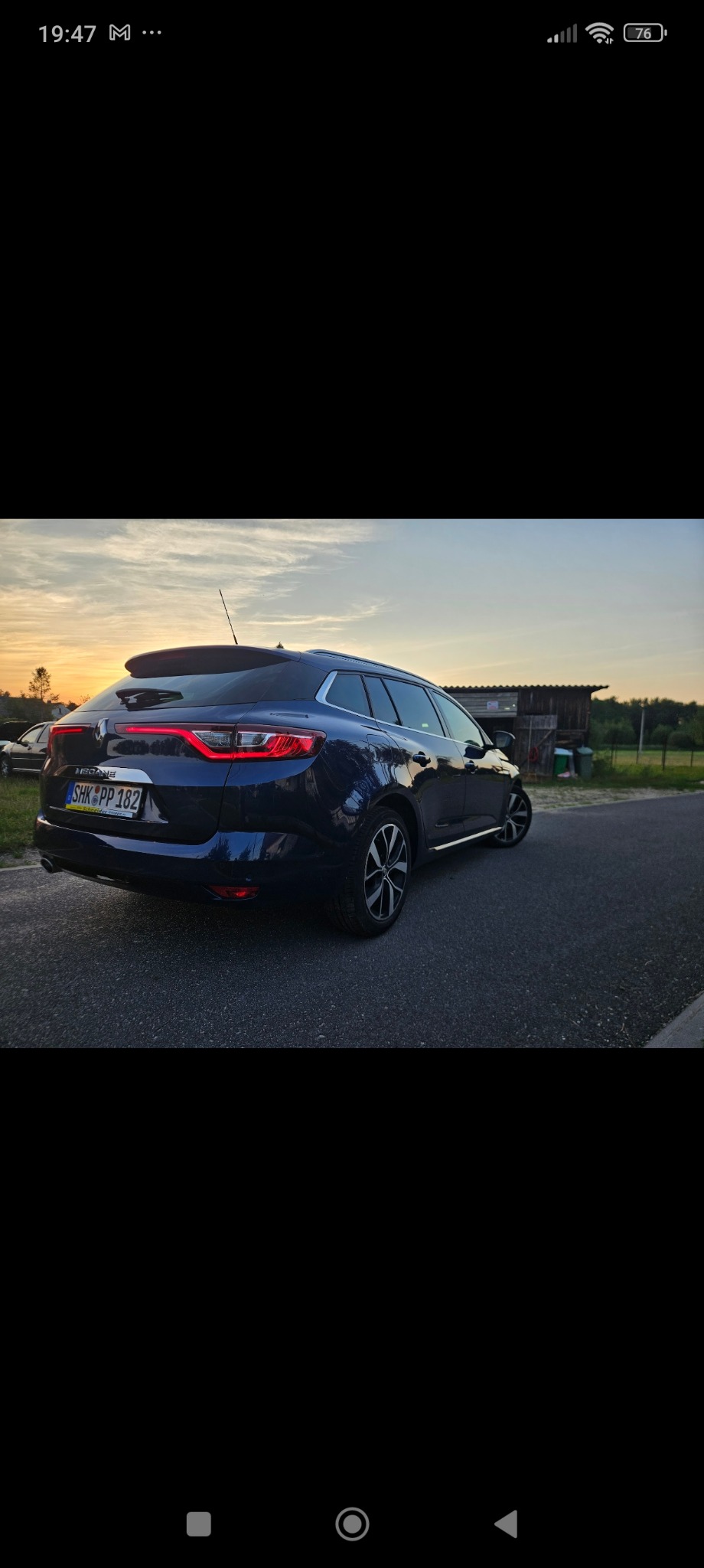 Granatowy samochód kombi Renault Megane, zaparkowany na asfalcie, widok z tyłu, w tle zachód słońca i budynek gospodarczy.