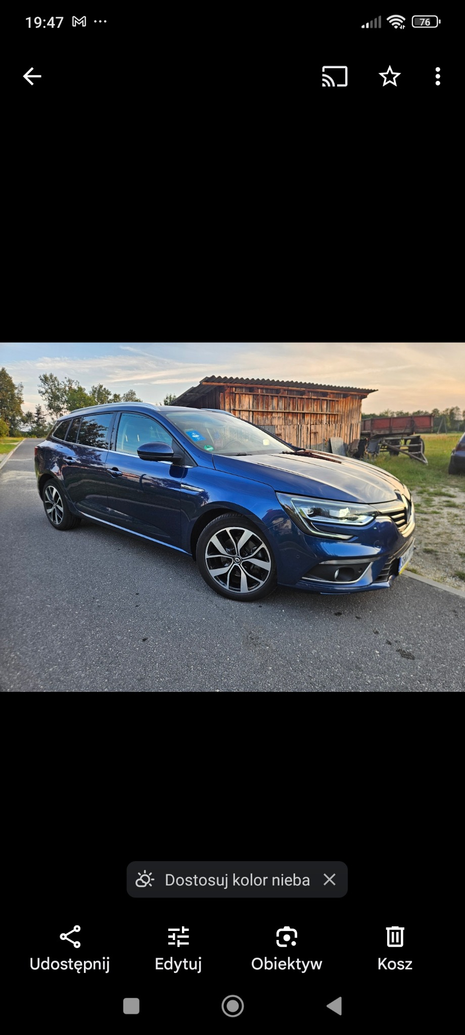 Niebieski samochód kombi marki Renault zaparkowany na asfaltowej drodze, z widocznym budynkiem gospodarczym w tle.