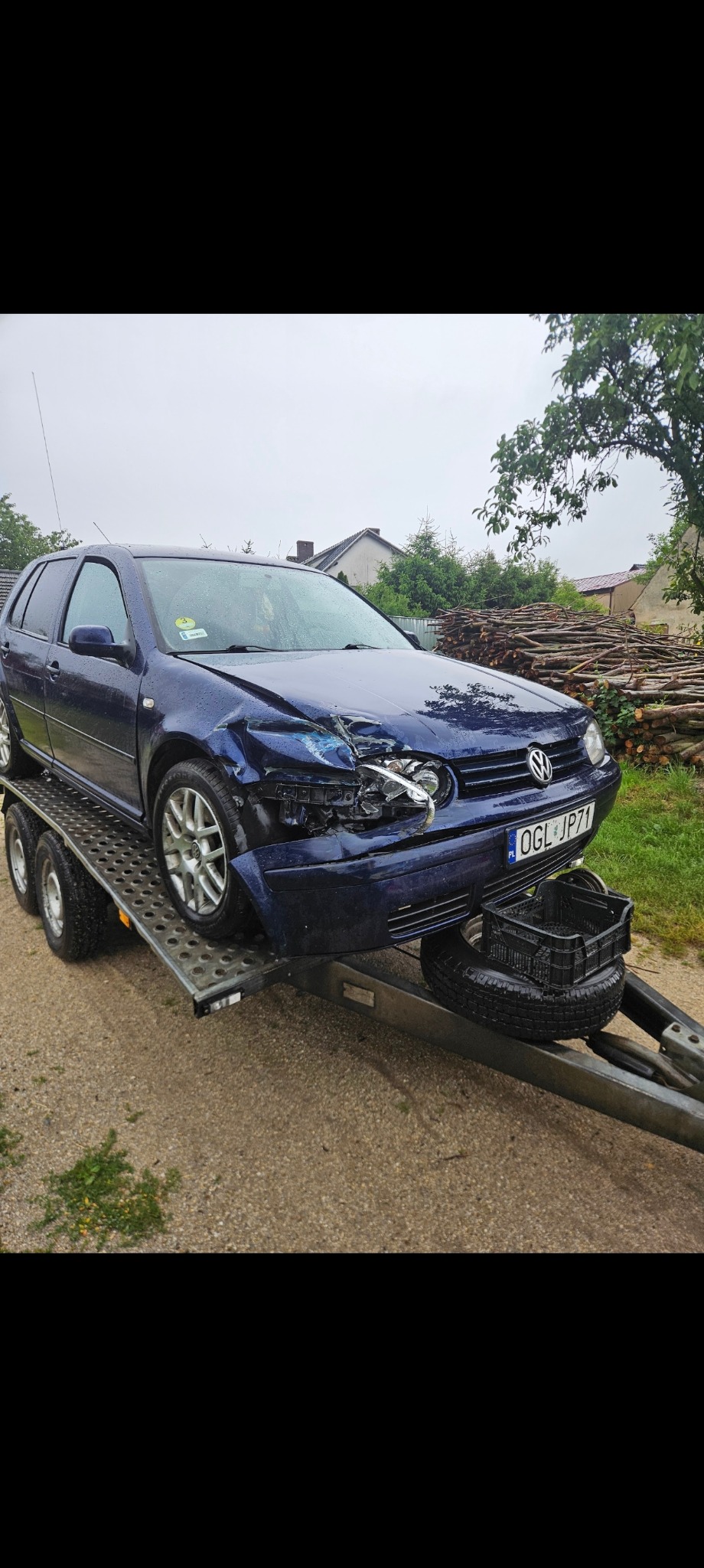 Uszkodzony, granatowy Volkswagen Golf na lawecie, z widocznymi wgnieceniami i uszkodzeniami reflektora po stronie kierowcy, tablica rejestracyjna OGL JP71, w tle sterta drewna.