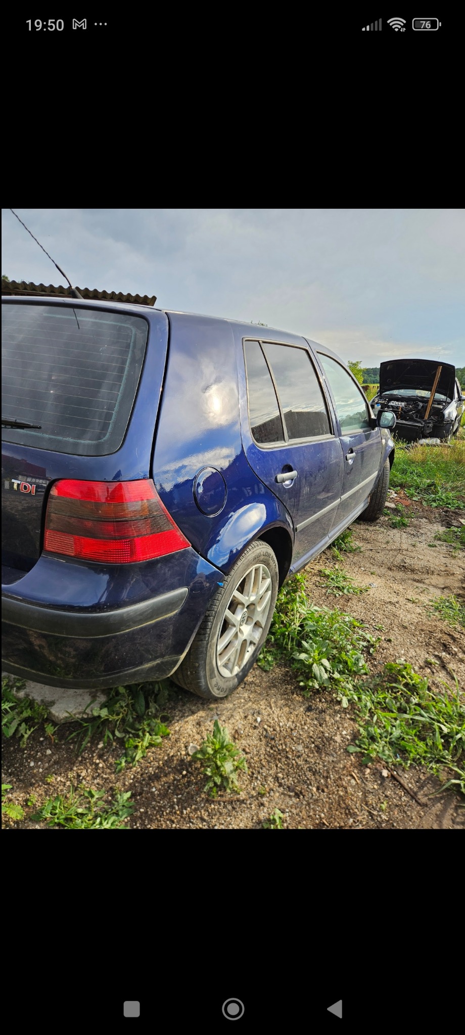 Błękitny Volkswagen Golf TDI z widocznym uszkodzeniem lakieru na tylnym błotniku, stojący na nierównym terenie obok drugiego samochodu z otwartą maską.