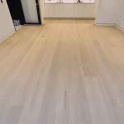 Jasne, szerokie panele podłogowe w nowoczesnym wnętrzu z białą zabudową kuchenną i czarną ramą drzwiową, widoczne z perspektywy podłogi