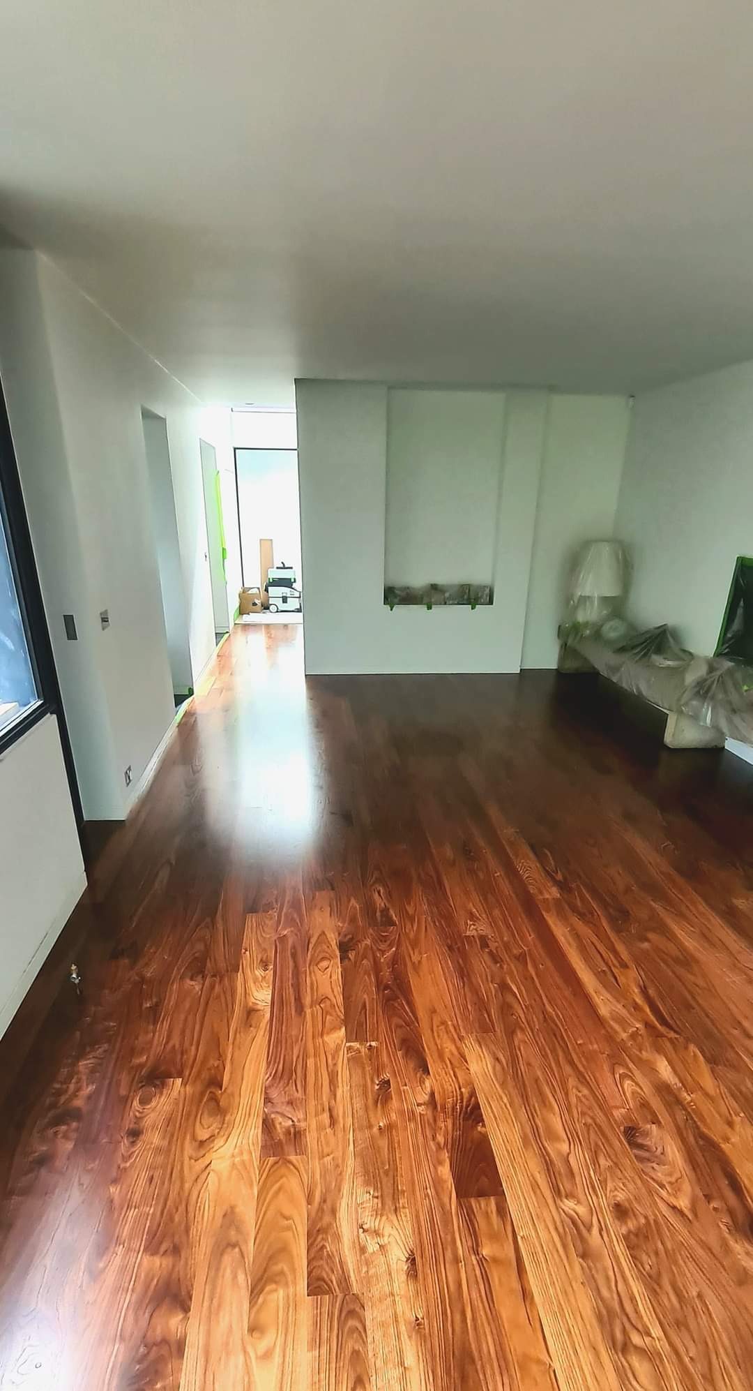 Wnętrze z nowo ułożoną, błyszczącą podłogą z ciemnego drewna, widok na dalszą część mieszkania z meblami zabezpieczonymi folią malarską.