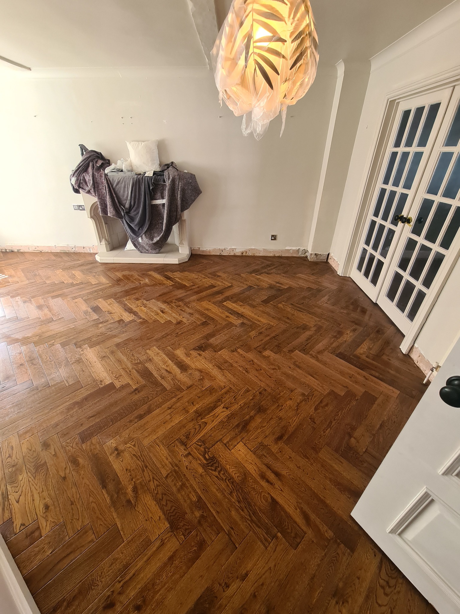 Ukończona podłoga z parkietu ułożonego w jodełkę w pomieszczeniu z białymi ścianami, kominkiem i podwójnymi drzwiami balkonowymi, widoczne ślady po listwach przypodłogowych.