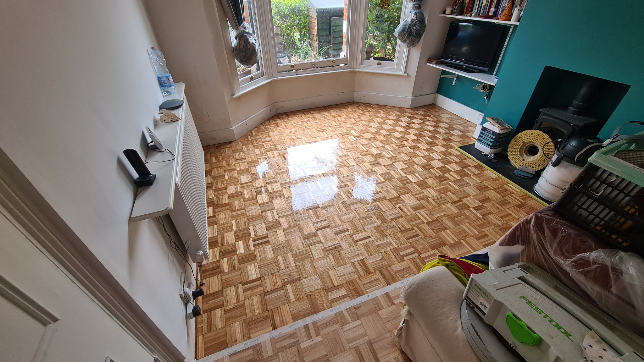 Parkiet z małych kwadratów po cyklinowaniu, z widocznym połyskiem i odbiciami światła, narzędzia do cyklinowania w rogu pokoju, okno wykuszowe z widokiem na ogród.