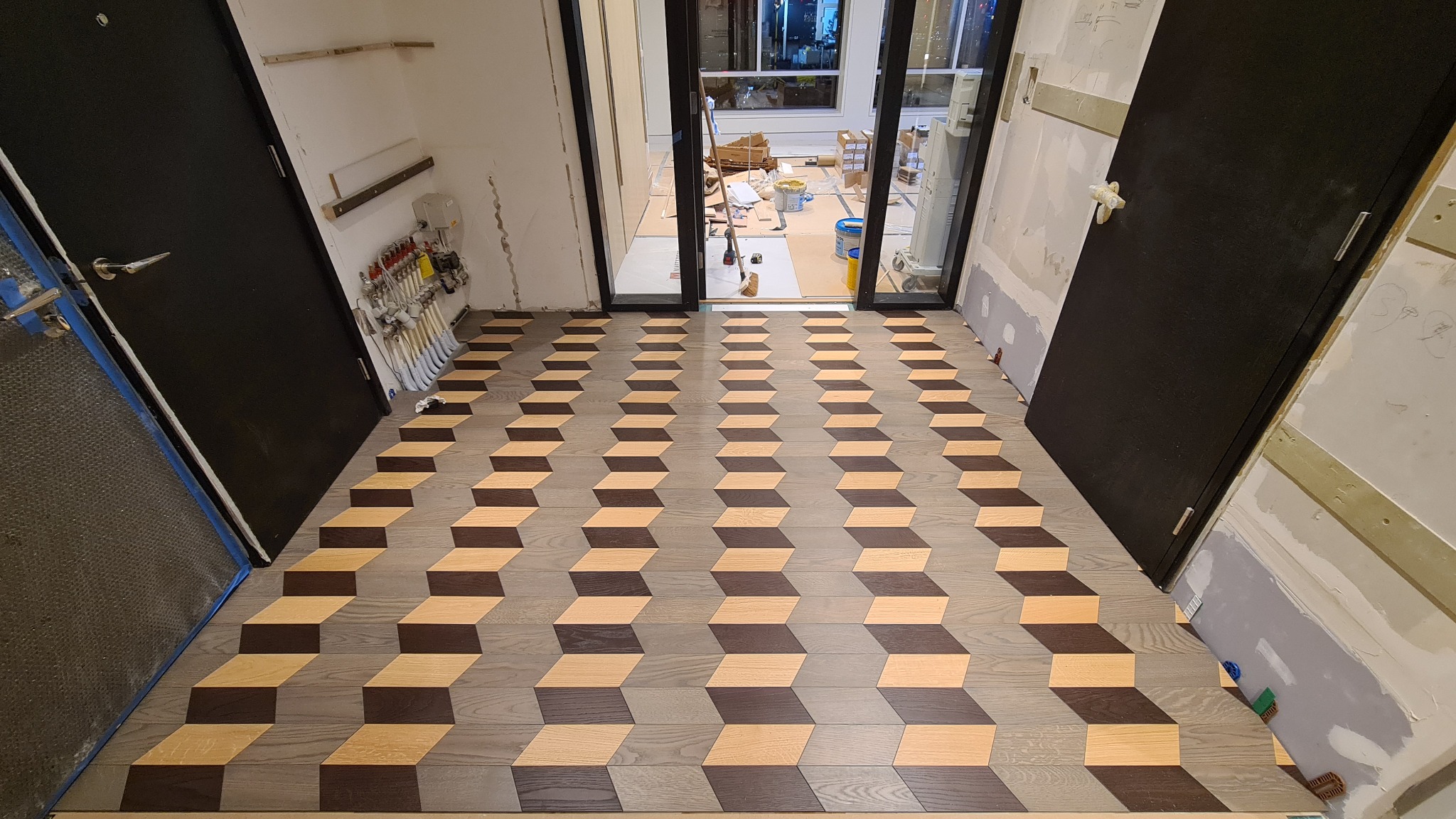 Ukończona podłoga z paneli ułożonych we wzór 3D, widoczne dwie czarne drzwi i przygotowane ściany do dalszych prac wykończeniowych, w tle widok na miasto przez szklane drzwi.