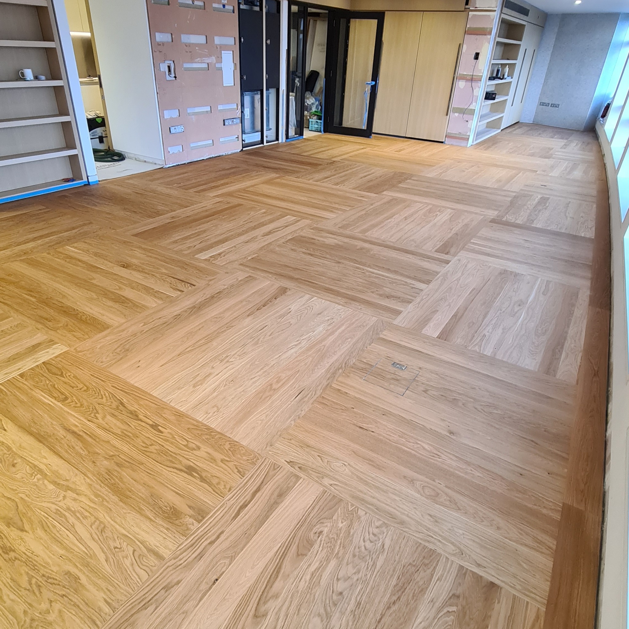 Nowo ułożony parkiet w jodełkę francuską z widocznym włóknem drewna, z elementami wykończeniowymi i gniazdem podłogowym, w pomieszczeniu w trakcie remontu.