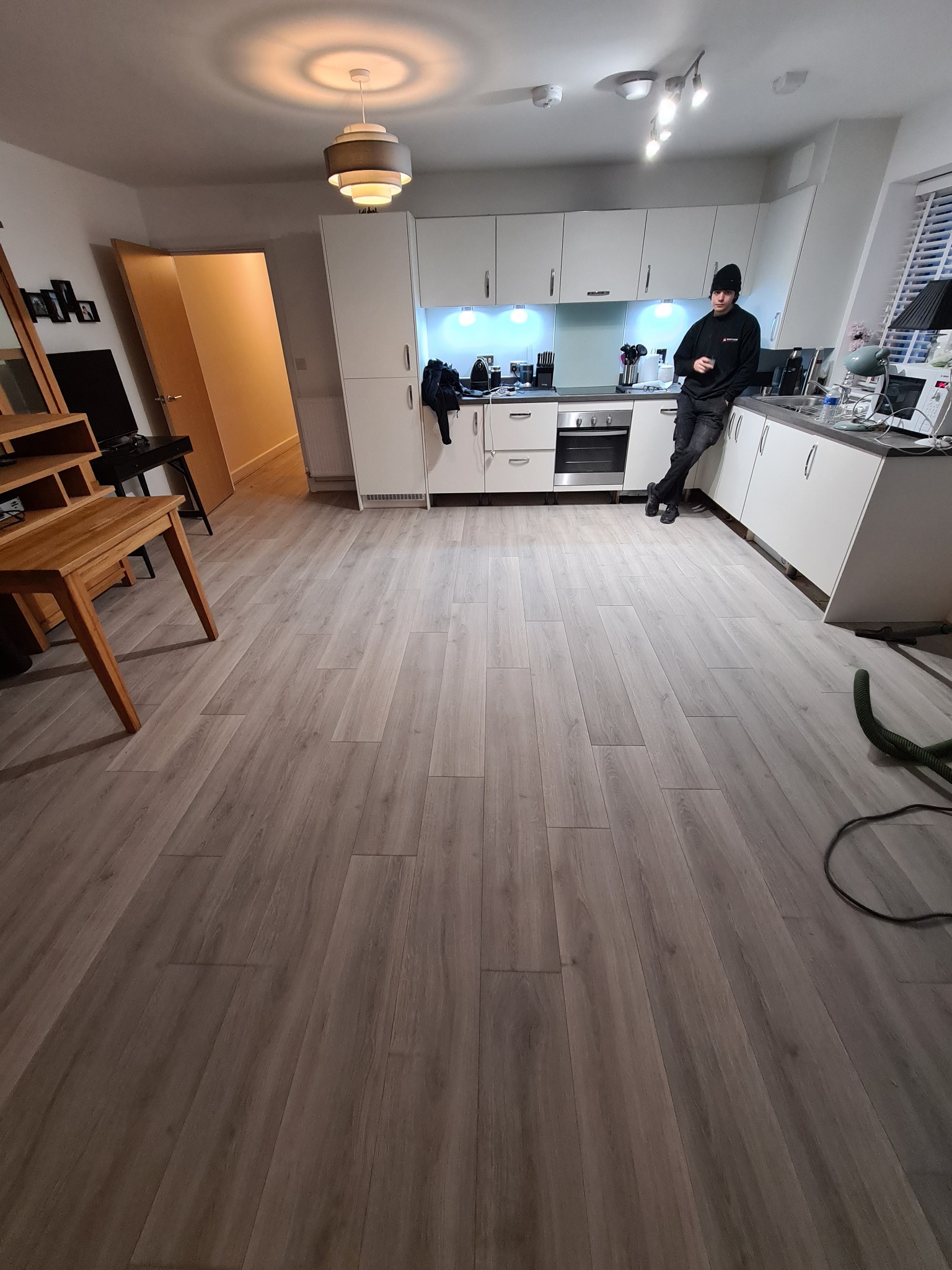 Jasnoszare panele podłogowe w kuchni, widoczny fragment węża od odkurzacza oraz mężczyzna oparty o blat kuchenny.
