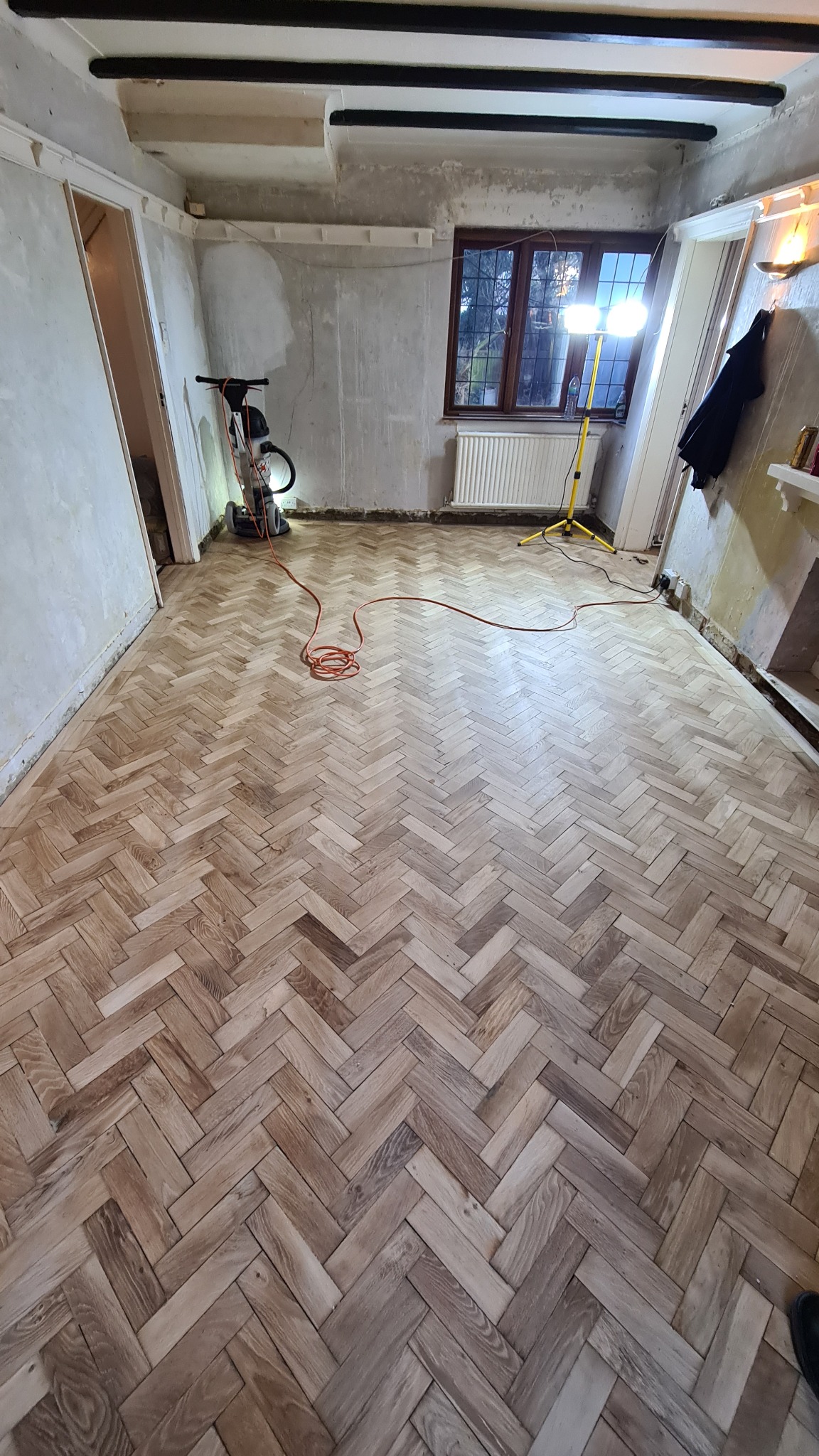 Parkiet ułożony w jodełkę podczas renowacji, widoczna maszyna do cyklinowania i lampa budowlana oświetlająca pomieszczenie, ściany w stanie surowym.