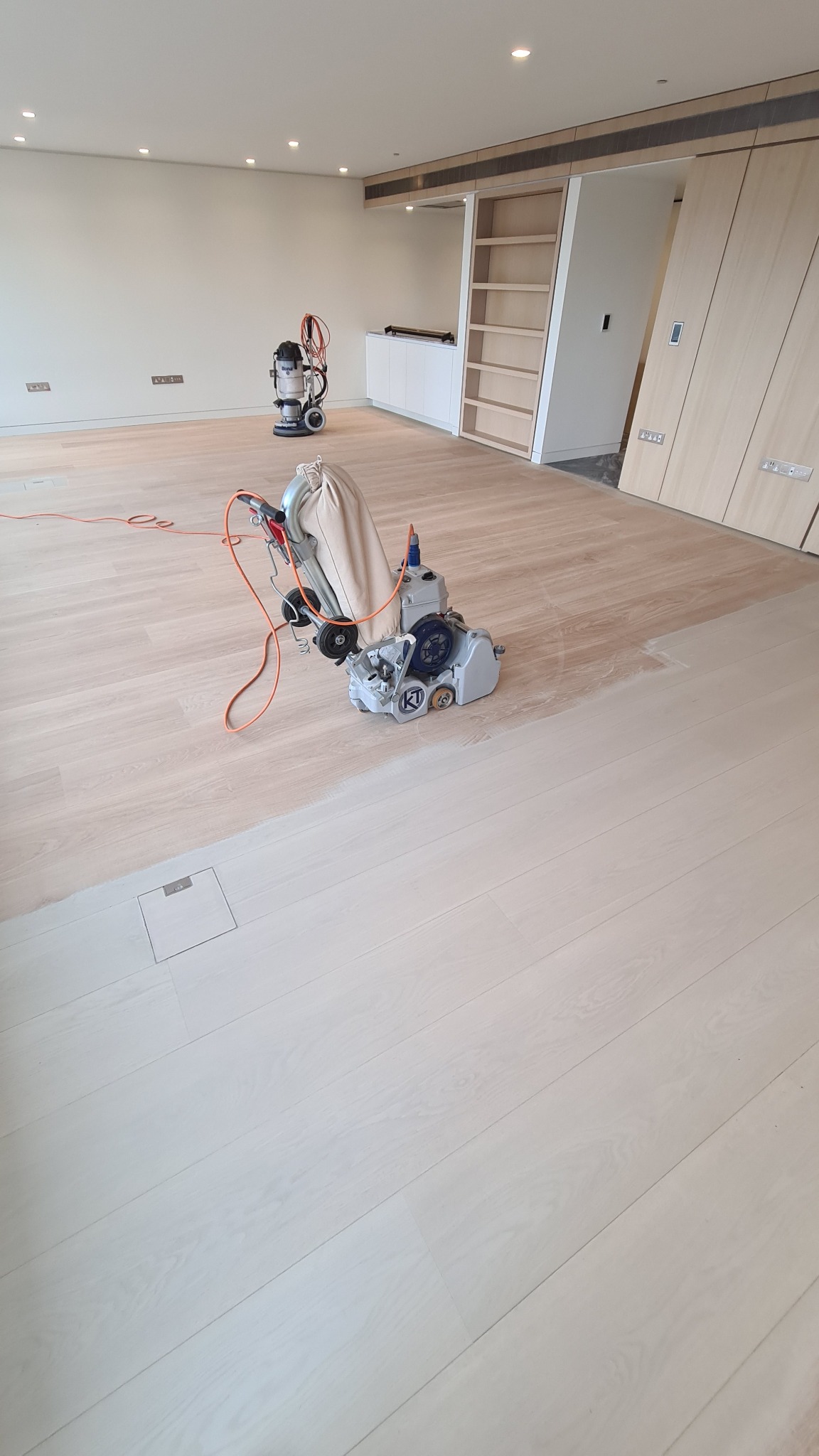 Parkiet podczas cyklinowania; widoczna maszyna do cyklinowania z workiem na pył i odkurzacz przemysłowy w tle, część podłogi już wycyklinowana, ukazująca jaśniejszy kolor drewna.