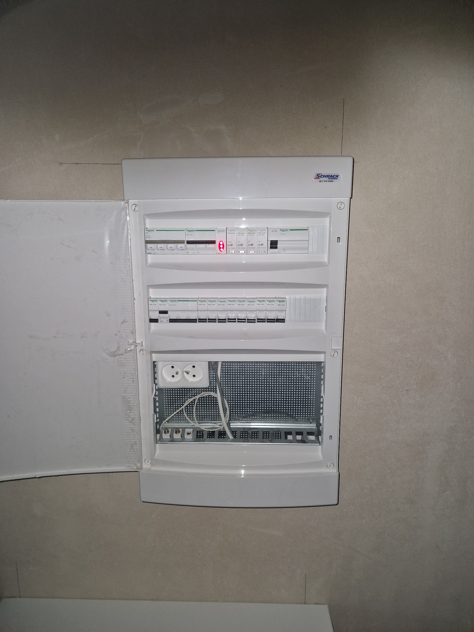 Rozdzielnica elektryczna Schrack Technik z otwartymi drzwiczkami, widoczne zabezpieczenia, gniazda i okablowanie. Ściana w tle o szarej, surowej fakturze.