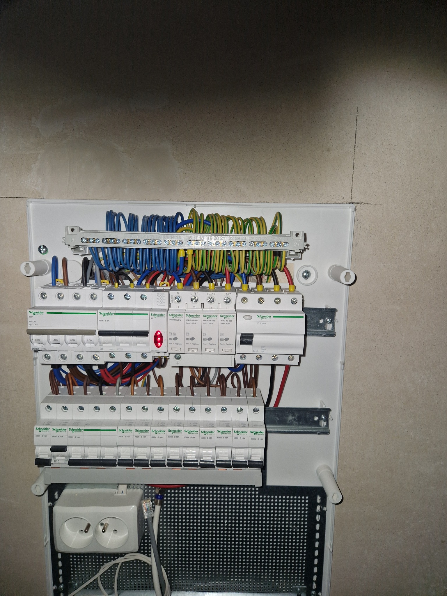 Rozdzielnica elektryczna Schneider z zainstalowanymi bezpiecznikami i okablowaniem, widoczna na tle surowej ściany. Dwa gniazdka elektryczne w dolnej części.