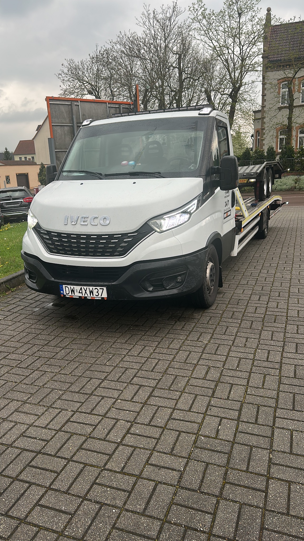 Biała laweta Iveco na brukowanym placu, tablica rejestracyjna DW 4XW37. Widok na przednią część pojazdu i platformę do transportu. Budynek w tle.