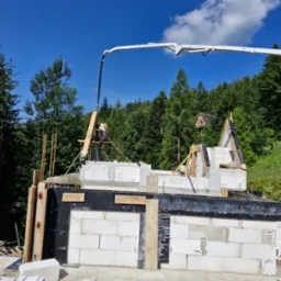 Budowa domu jednorodzinnego: wznoszenie ścian z bloczków betonowych, widoczny pracownik na rusztowaniu oraz pompa do betonu z wysięgnikiem, w tle las i fragment dachu innego budynku.