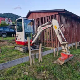 Mini koparka Takeuchi TB216 na trawiastym terenie budowy, obok drewnianego szalunku fundamentów i szopy z pionowych desek, w tle zabudowa wiejska.