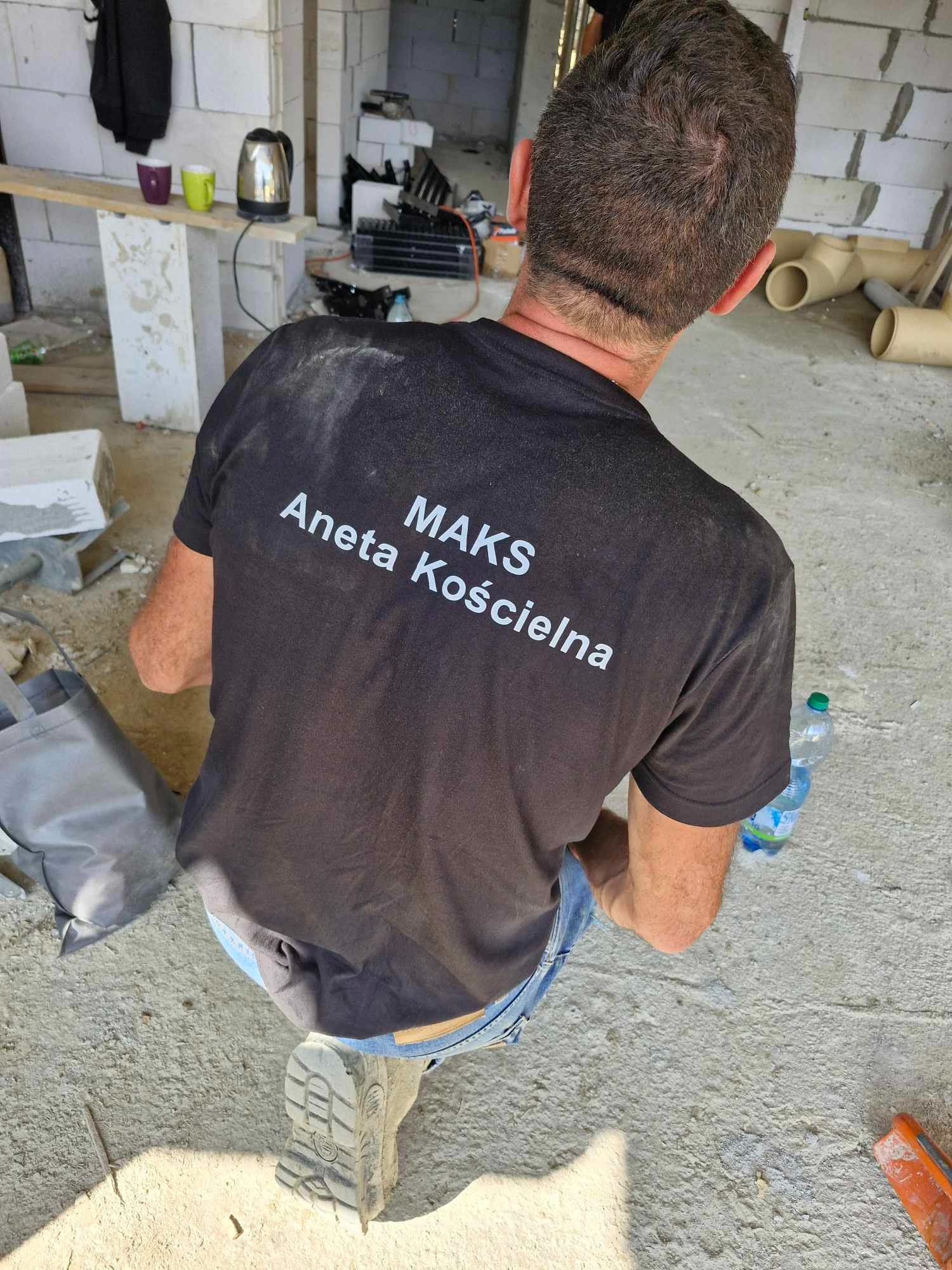 Murarz w czarnej koszulce z nadrukiem 'MAKS Aneta Kościelna' klęczy na betonie w trakcie pracy, w tle widać niedokończone ściany z białych bloczków, rury wentylacyjne i prowizoryczny stół...