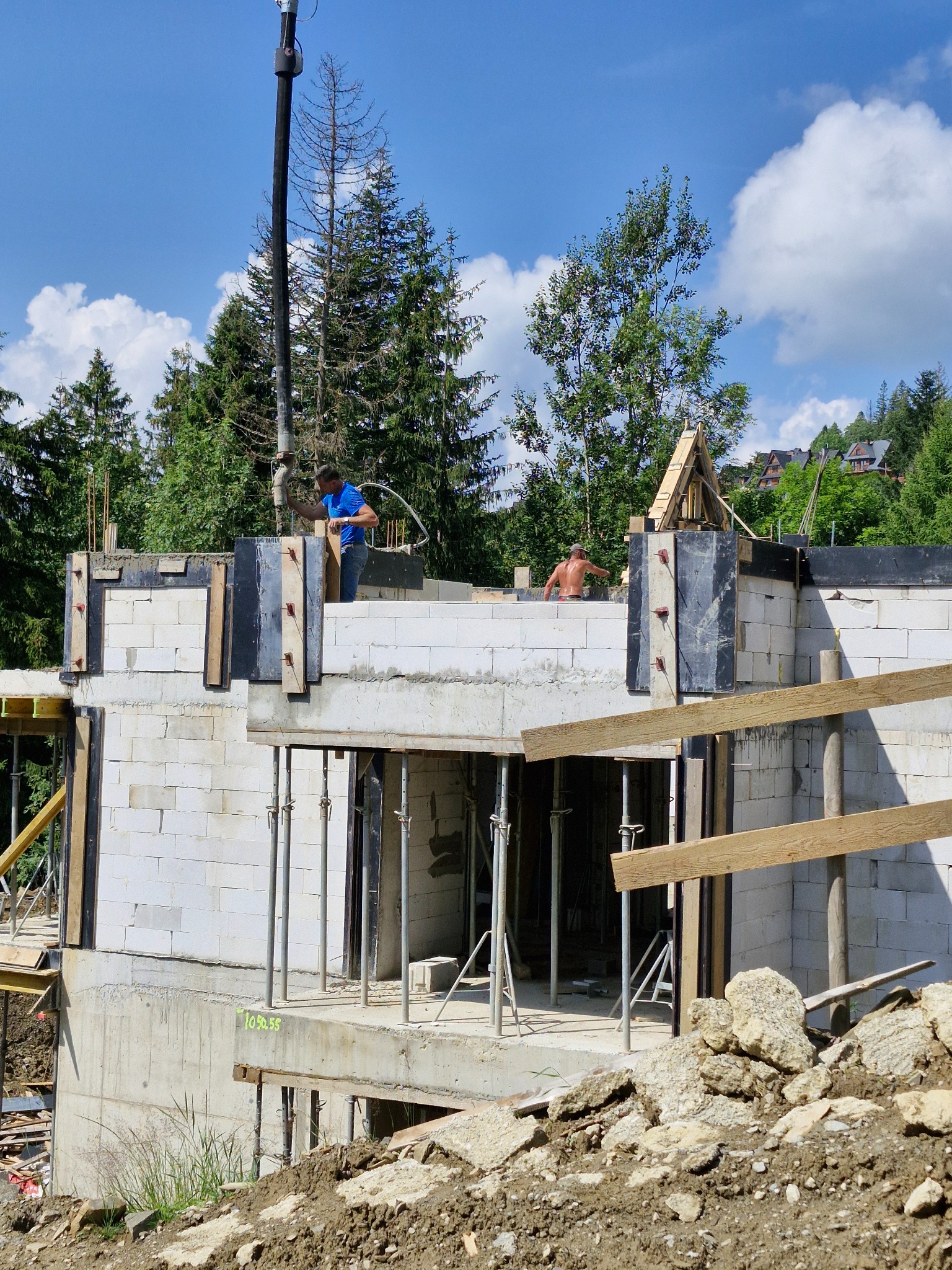Budowa domu w górach: wylewanie betonu na ściany z białych bloczków, widoczny pracownik w niebieskiej koszulce i drugi bez koszulki, las w tle, błękitne niebo z chmurami.