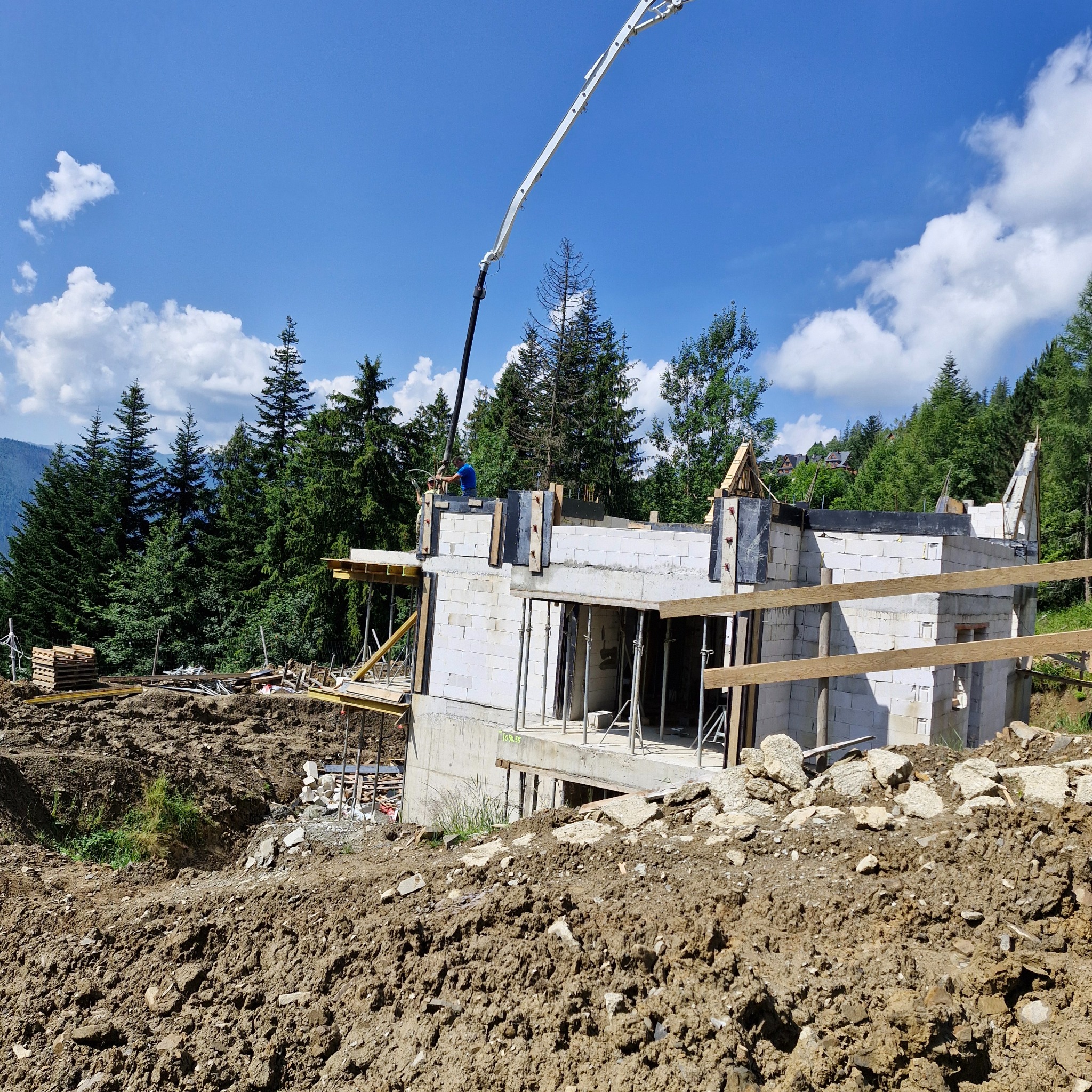 Budowa domu murowanego w górskim otoczeniu, widoczna pompa do betonu, ściany z bloczków, drewniane szalunki i zbrojenia, w tle las iglasty pod błękitnym niebem.