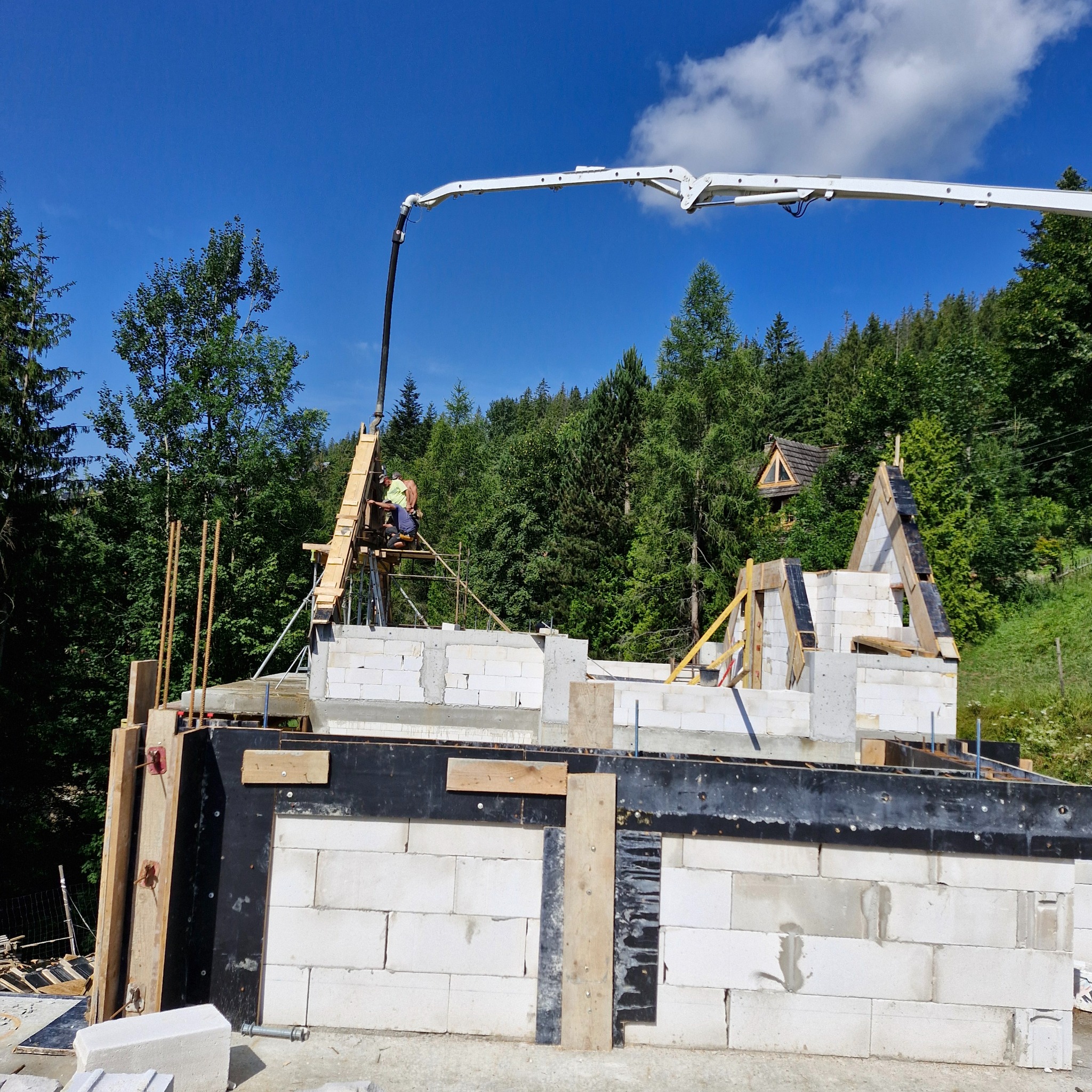 Budowa domu jednorodzinnego: wznoszenie ścian z bloczków betonowych, widoczny pracownik na rusztowaniu oraz pompa do betonu z wysięgnikiem, w tle las i fragment dachu innego budynku.