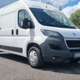 Biały Peugeot Boxer na parkingu, widok z przodu, lekki cień, drzewa i słupy oświetleniowe w tle.