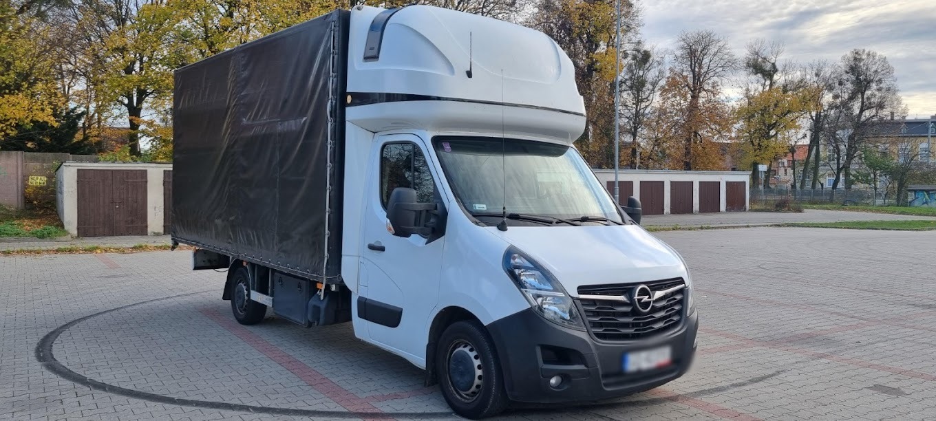 Biały samochód dostawczy Opel z czarną plandeką na parkingu z kostki brukowej, w tle budynki i drzewa z jesiennymi liśćmi.