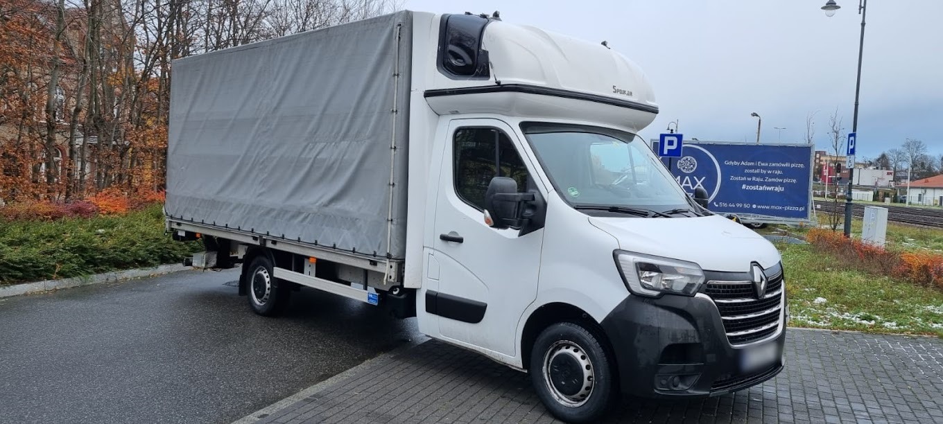 Biały samochód dostawczy Renault Master z wysoką kabiną i plandeką, zaparkowany na parkingu, z widocznym billboardem reklamowym w tle.