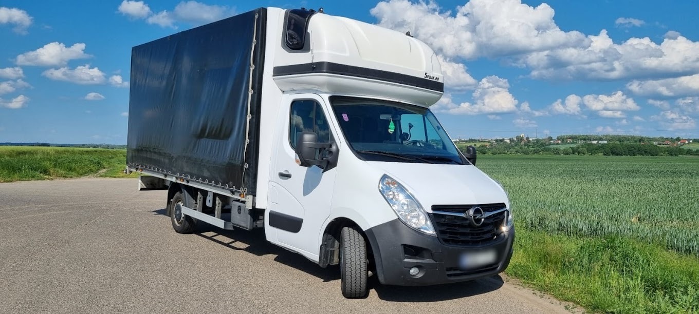 Biały samochód dostawczy z plandeką marki Opel Master, zaparkowany na asfaltowej drodze z widokiem na zielone pola i błękitne niebo z chmurami.