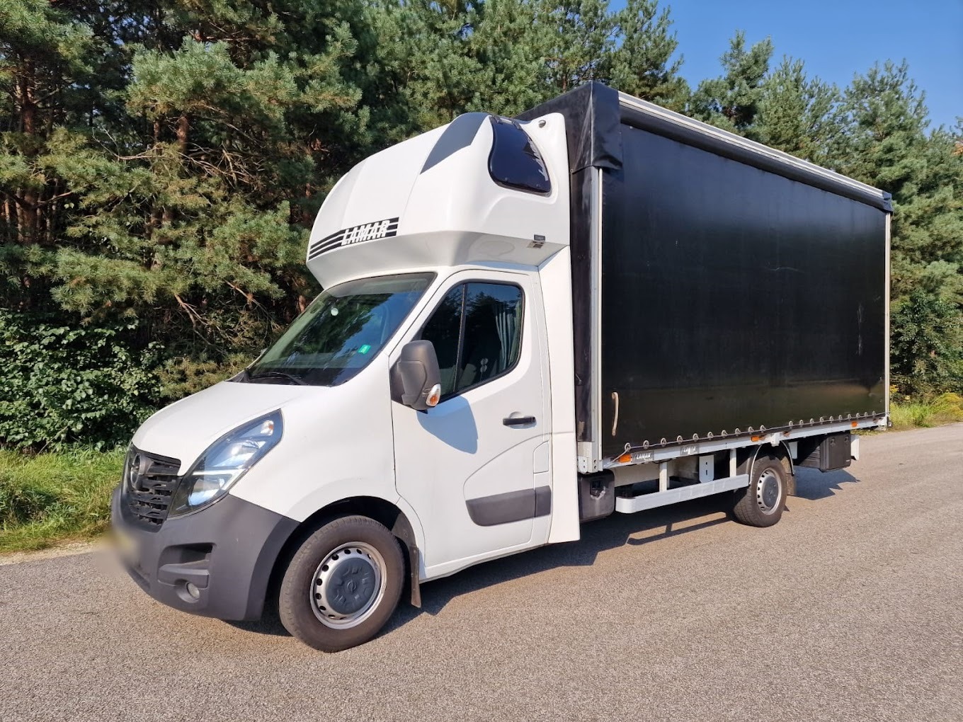 Biały samochód dostawczy z czarną plandeką na tle drzew, zaparkowany na asfaltowej drodze w słoneczny dzień.