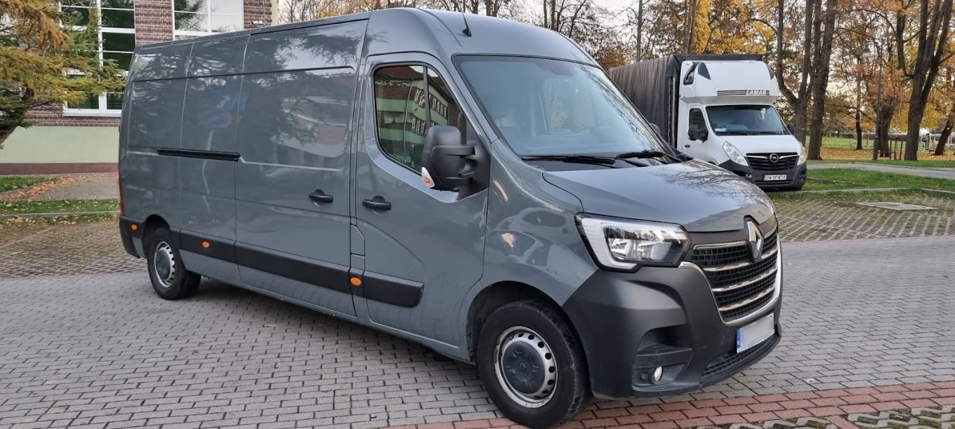 Szary Renault Master zaparkowany na kostce brukowej, widoczny z boku, z innym busem w tle po prawej stronie.