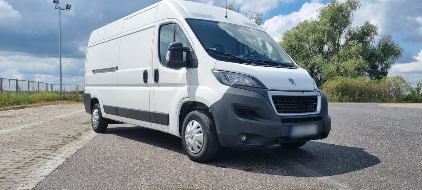 Biały Peugeot Boxer na parkingu, widok z przodu, lekki cień, drzewa i słupy oświetleniowe w tle.