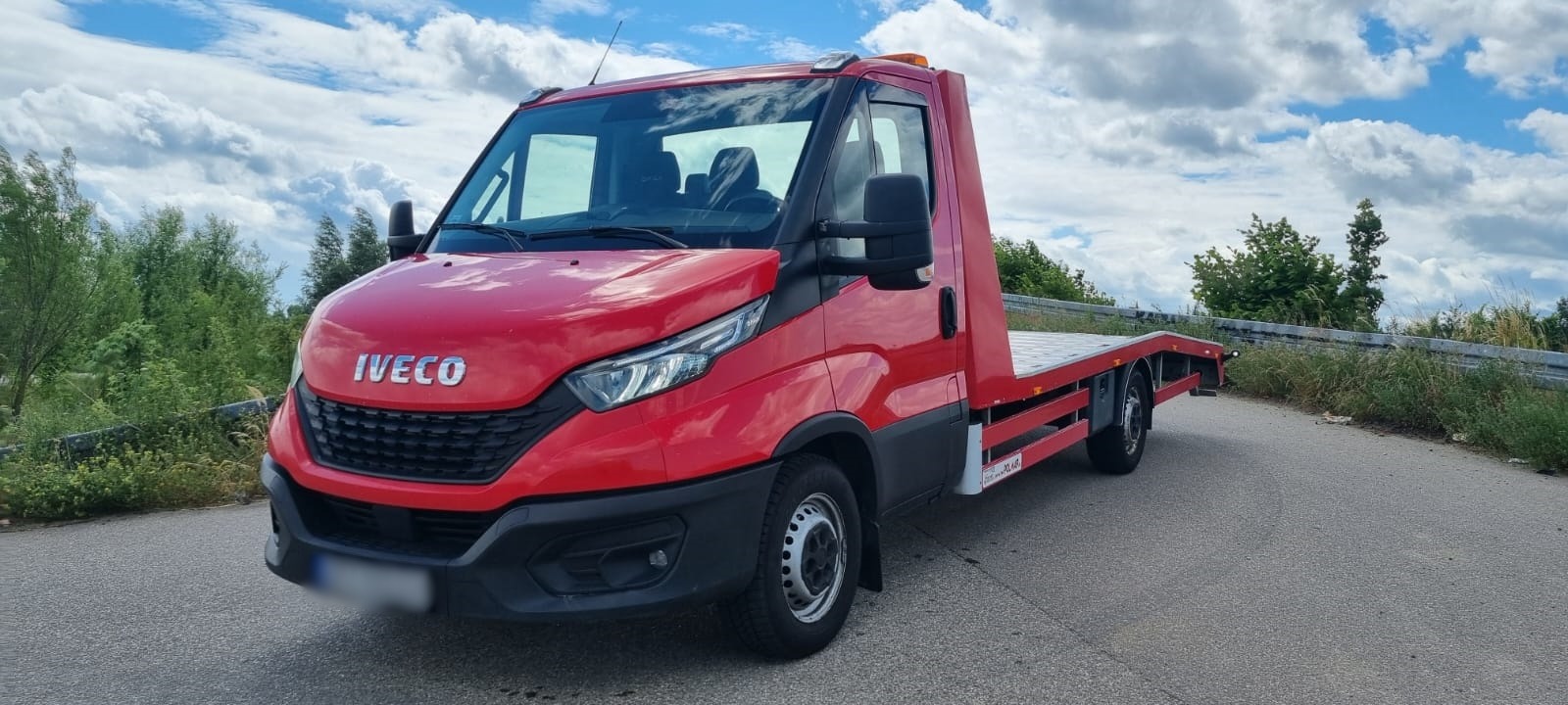 Czerwona laweta Iveco na asfalcie w słoneczny dzień. Widoczny pusty najazd i światła ostrzegawcze. Tło stanowią drzewa i błękitne niebo z chmurami.