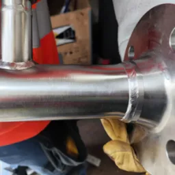 Spoina na rurze ze stali nierdzewnej, widoczne ślady obróbki i rękawice robocze w tle.