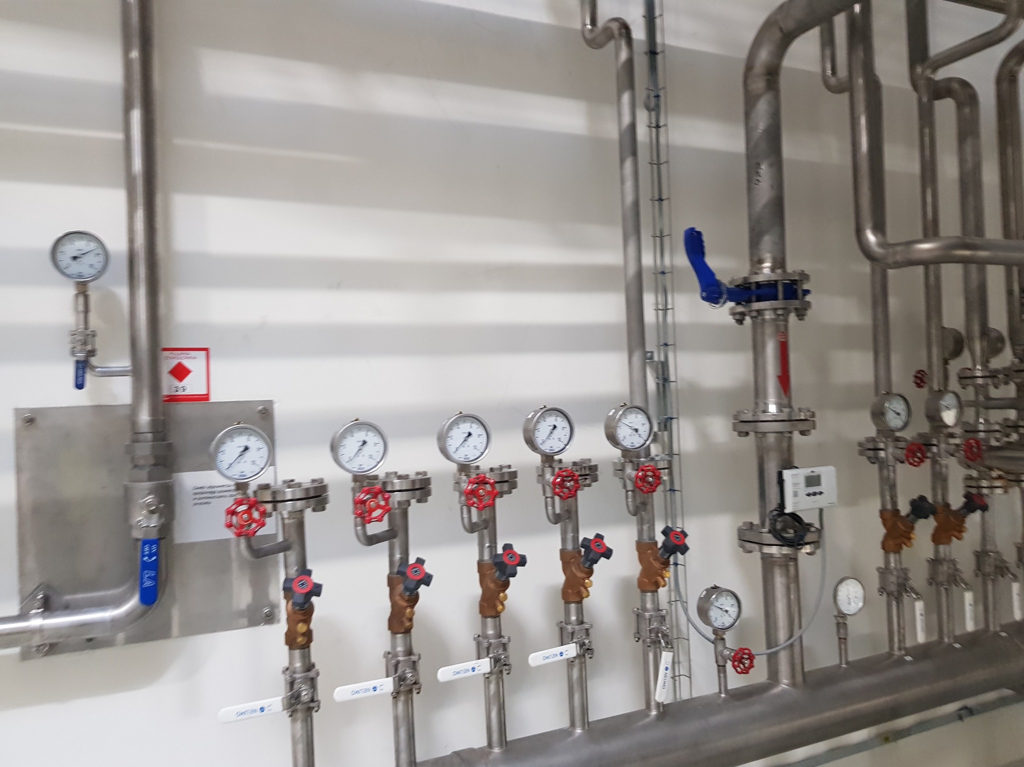 Instalacja przemysłowa z rzędami rur ze stali nierdzewnej, zaworami z czerwonymi pokrętłami i manometrami na białej ścianie.
