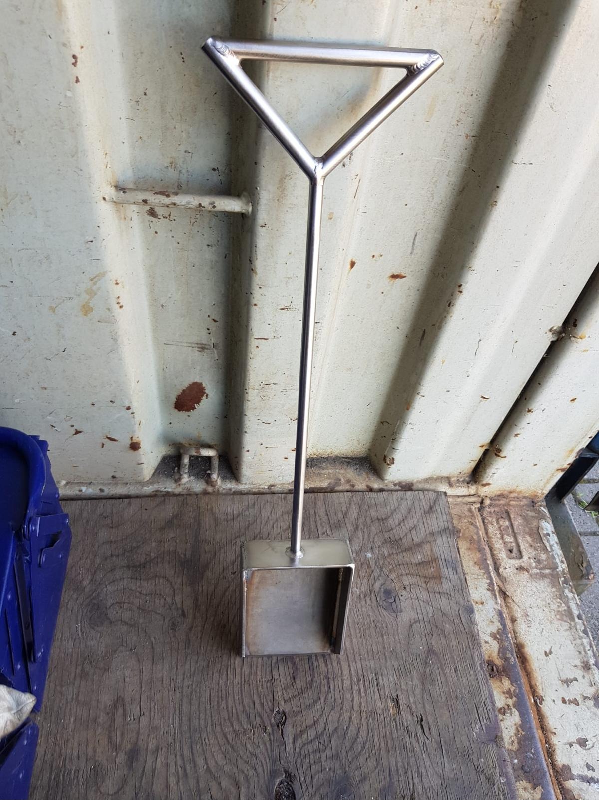 Metalowa szufelka z uchwytem w kształcie trójkąta, wykonana z polerowanej stali, leżąca na drewnianej powierzchni przy ścianie kontenera.