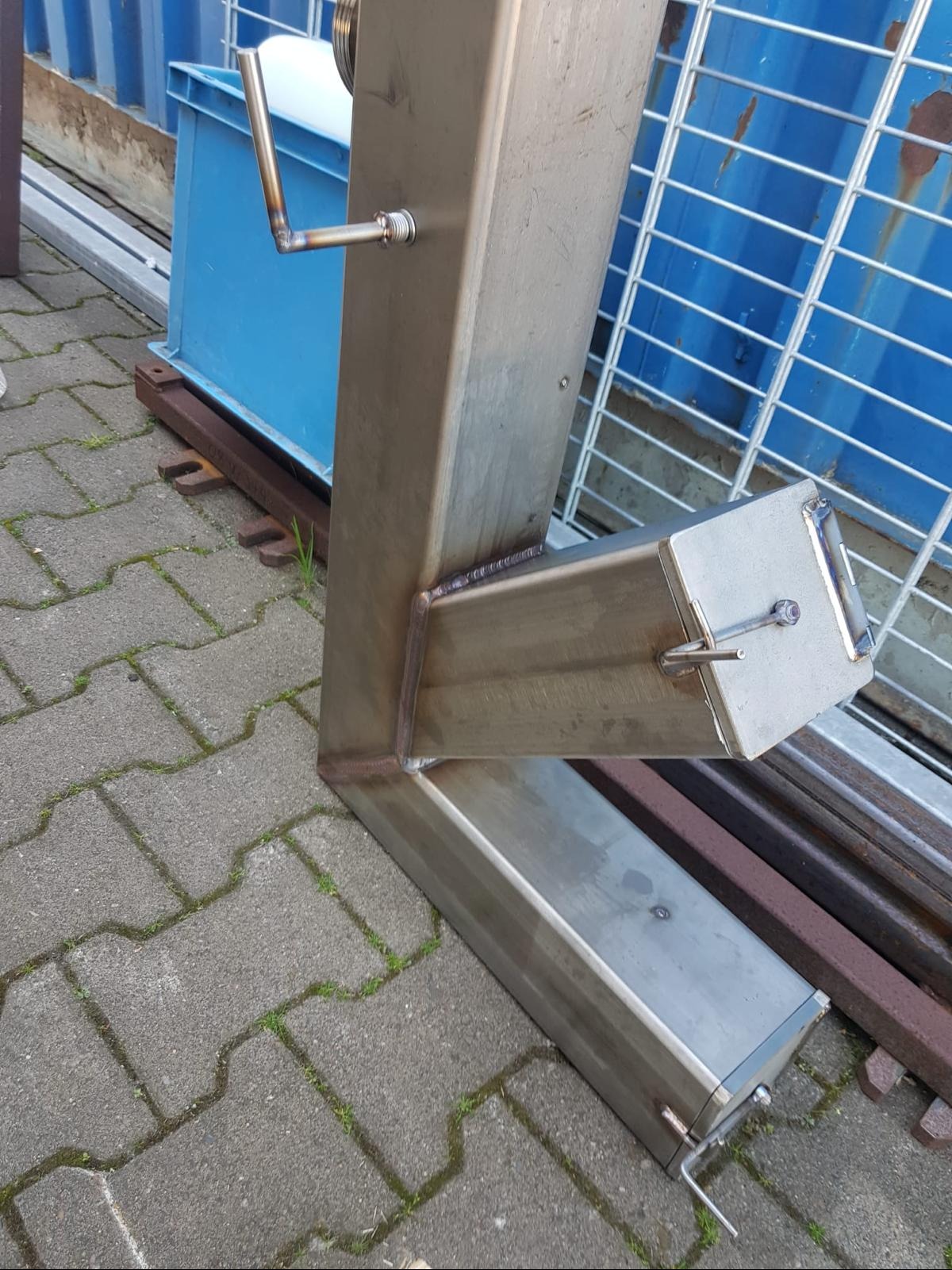 Metalowa konstrukcja o profilu litery L, wykonana ze stali nierdzewnej, z widocznymi spawami, stojąca na brukowanej powierzchni, z niebieskim kontenerem w tle.