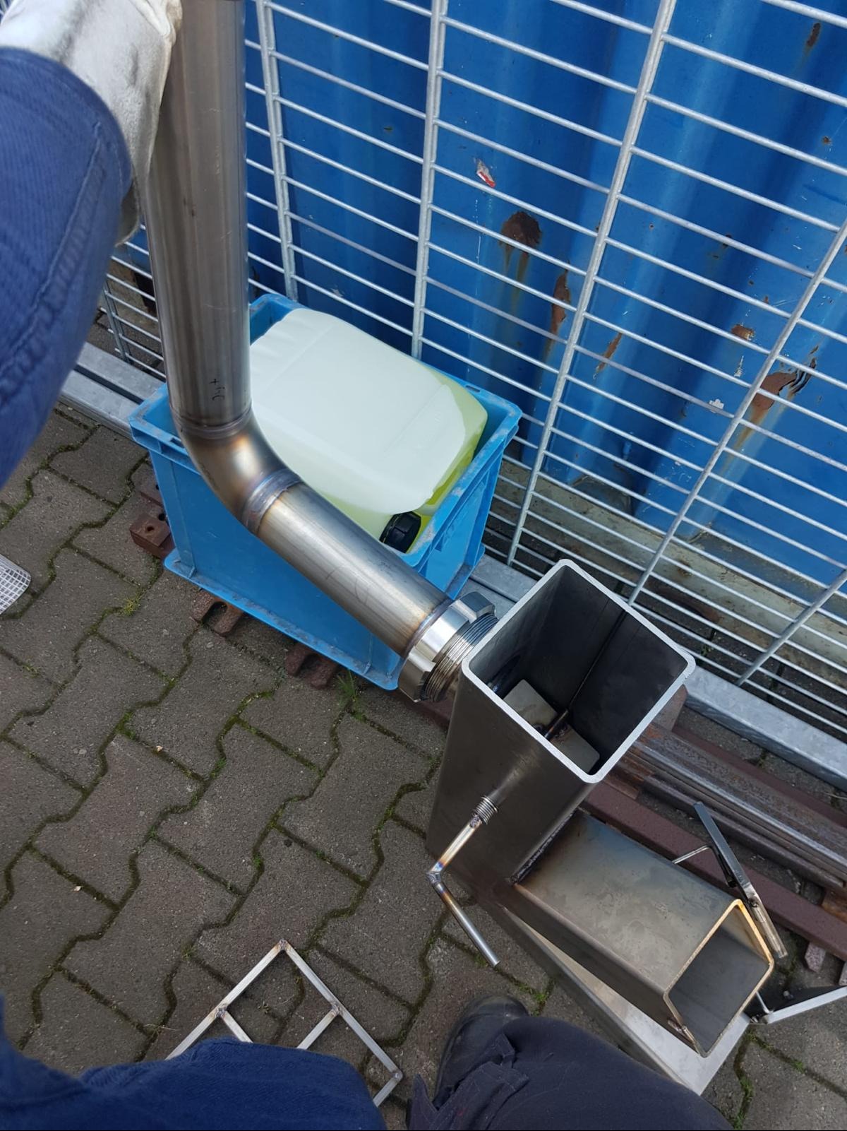 Połączenie spawane rury okrągłej z profilem kwadratowym, widoczne ślady spawania, niebieski pojemnik i metalowa krata w tle.