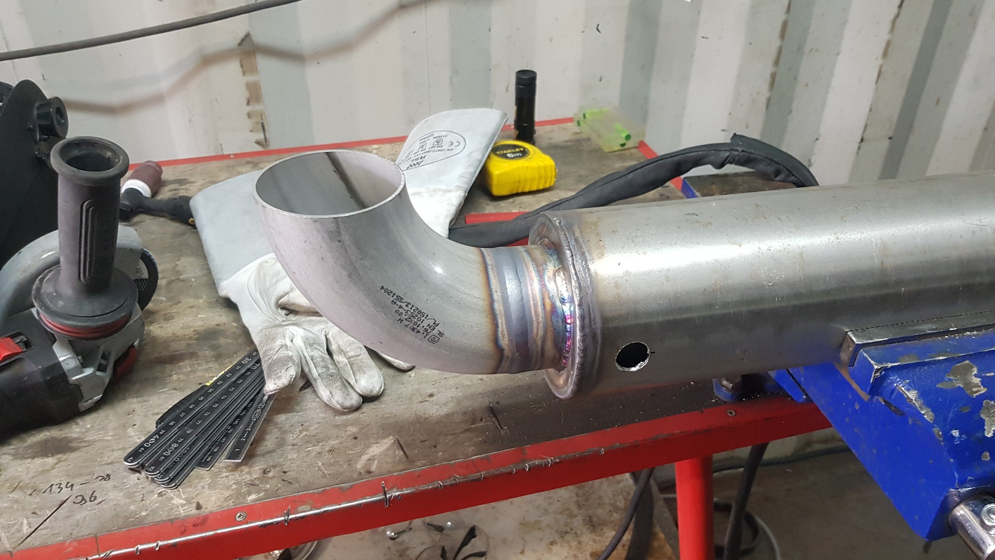 Spoina rury ze stali nierdzewnej z widocznymi kolorami hartowania, obok leżą rękawice ochronne i miarka zwijana.