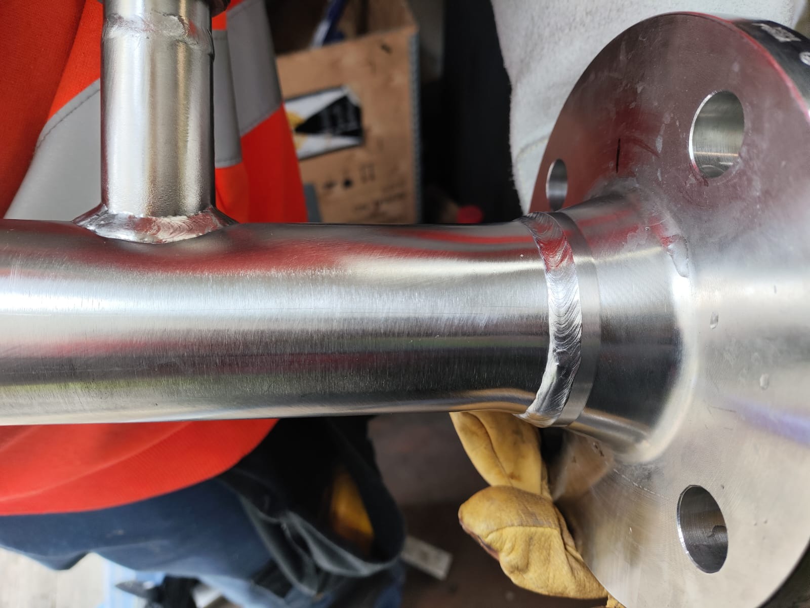 Zespawany element rurociągu ze stali nierdzewnej z widocznymi śladami po spawaniu, trzymany w rękawicach ochronnych, z widocznym kołnierzem i spawem TIG.