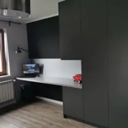 Ciemnoszara zabudowa meblowa z biurkiem i szafą w pokoju, minimalistyczny design, lampka ścienna, częściowo widoczne okno, zabawka - czerwony samochód.