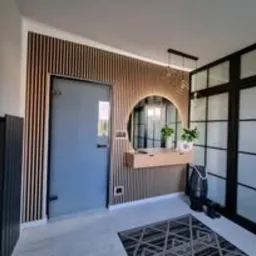 Nowoczesny przedpokój: szara ściana z lamelami, okrągłe lustro z podświetleniem LED, drewniana konsola i czarna, przeszklona ściana działowa.