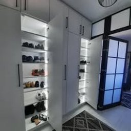 Meble Wręczycki Paweł Wręczycki - Biała szafa na wymiar z oświetleniem LED i otwartymi drzwiami, ukazująca półki z butami. Widoczny fragment dywanu i drzwi z czarnymi ramami.