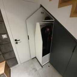 Meble Wręczycki Paweł Wręczycki - Szafa wnękowa pod schodami z białymi frontami, otwarta, ukazująca przechowywane przedmioty. Ściana z szare panele dekoracyjne, białe drzwi po lewej.