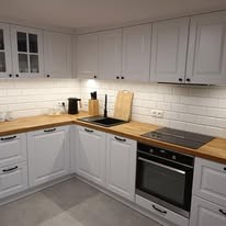 Biała kuchnia z drewnianym blatem i czarnymi uchwytami, widok z niskiego kąta, płytki metro na ścianie, czarny zlew i płyta indukcyjna.