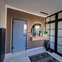 Nowoczesny przedpokój: szara ściana z lamelami, okrągłe lustro z podświetleniem LED, drewniana konsola i czarna, przeszklona ściana działowa.