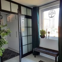 Biała szafa wnękowa z czarnymi profilami i matowym szkłem, widoczna z boku, z dekoracjami okiennymi i rośliną w tle.