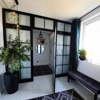 Nowoczesna szafa wnękowa z czarnymi ramami i matowym szkłem, dzieląca przestrzeń. Widoczny fragment wnętrza z lustrem i rośliną w donicy. Oświetlenie dekoracyjne.