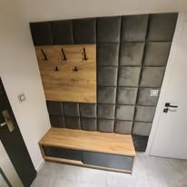 Tapicerowany panel ścienny z siedziskiem i wieszakami w przedpokoju. Drewno w odcieniach brązu kontrastuje z szarością tapicerki.
