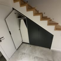 Biało-grafitowa szafa pod schodami z otwartymi drzwiami, ukazująca półki i deskę do prasowania. Drewniane schody na białej ścianie, szara podłoga.