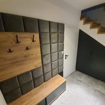 Nowoczesny przedpokój z panelem tapicerowanym w kratę, drewnianym wieszakiem i siedziskiem. Schody z drewnianymi stopniami w tle.