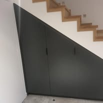 Szara szafa wnękowa pod schodami z drewnianymi stopniami, minimalistyczny design, białe ściany, widok z dołu.